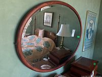 Front angle view of round wood framed mirror hanging on green wall with bedroom furniture reflected.