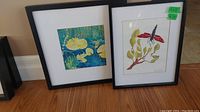 Two framed monotype prints, one with blue and yellow floral design and the other with red flower and green leaves, leaning against wall.