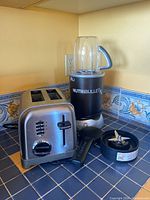 Photo showing the NutriBullet RX blender with blending cup on the base, blender blade assembly, tamper tool, and a stainless steel 4-slice toaster on a tiled kitchen counter.