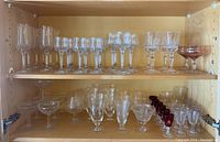 Photo showing upper and lower shelves of clear and ruby stemmed glasses of various styles and sizes arranged inside a wooden cabinet.