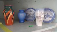 Four decorative items on a shelf: two blue and white Royal Copenhagen plates with windmill and countryside scenes, one white Royal Copenhagen vase with blue countryside design, one solid blue small vase, and one tall multicolor Bulgarian vase with abstract orange, black, and green design.