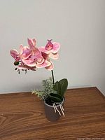 Front angle view of artificial pink orchid plant with green and white foliage in a brown pot on wooden surface.