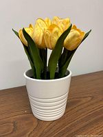 Side view of artificial yellow tulips in white ceramic vase placed on wooden surface, showing flower details and vase texture.