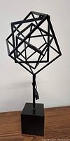 Front view of black metal geometric sphere composed of interlocking square frames mounted on a cube base.