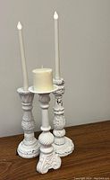Three matt white carved candle holders displayed on wooden surface. Two hold tall slender battery-operated faux candles, the third holds a small rustic gold pillar candle.