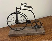 Side view of the old-fashioned bicycle metal sculpture showing large front wheel, smaller rear wheel, and wooden base.