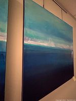 Side view of square canvas painting in blue and green hues, hanging on wall by chain.