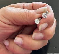 Hand holding two small stud earrings with clear cubic zirconia stones visible, showing size and detail of stones and setting.