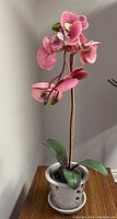 Front and top angled view of pink artificial orchid with multiple flowers and leaves in a gray ceramic pot on a wooden surface