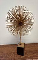 Full front view showing the gold star burst sculpture mounted on a black marble base against a white wall on wooden surface.