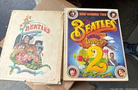 Photo showing two hardcover Beatles books by Alan Aldridge, 'The Beatles Illustrated Lyrics' and 'The Beatles Lyrics 2', placed side by side on a cardboard surface.