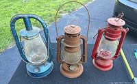 Photo of three kerosene lanterns standing outdoors on pavement, one blue, one brownish, one red.