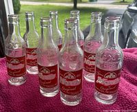 Photo of eight clear glass Moxie soda bottles with red and white labels, arranged on pink towel showing front labels and embossed details.