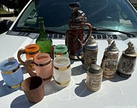 Overview of the lot showing all mugs, steins, decanter, green bottle, and wooden cup arranged on a white surface outdoors.