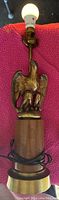 Full view of the lamp showing the brass eagle on wooden base with lightbulb on, cord coiled at bottom.
