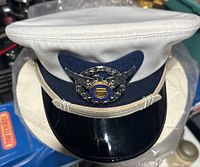 White peaked dress cap with glossy black visor and silver emblem with eagle and U.S. Coast Guard Auxiliary crest. Navy blue fabric band and silver hatcord visible. Hat protector cover partially visible beneath the hat.