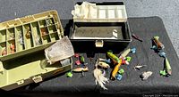 Two open plastic tackle boxes showing multiple small compartments containing various fishing lures and accessories, alongside about 20 loose lures arranged on a black surface.