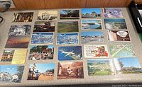 Top-down image showing all 25 postcards laid out in rows on a wooden floor, highlighting various scenic and historic views including Niagara Falls, airplanes, and tourist areas.