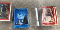 Photo showing three full sealed sets of E.T. The Extra-Terrestrial movie photo cards, the blue box labeled '87 Movie Photo Cards' and two cards with red film strip borders showing scenes from the movie featuring E.T. in costume.