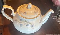 Photo of vintage Duchess Tranquility teapot, white porcelain with blue floral designs and gold trim from side angle showing spout and handle.