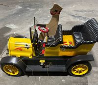 Side view of the yellow and black motorized toy car with driver figure and string control attached, showing detailed seats, wheels, and steering wheel.