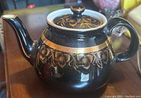 Side view of vintage Hall black ceramic teapot with gold floral vine decoration on the body and lid, showing the spout, handle and lid knob