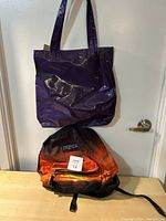 Photo of a purple shiny tote bag hanging above an orange and black Jansport backpack with a mountain sunset design.