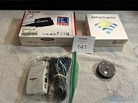 Top view showing BYTTEC USB hub box, Apple AirPort Express box, bundle of cables in plastic bag, and GE audio input switch device on a gray cloth surface.