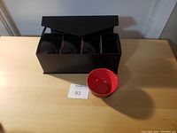 Four small red bowls arranged in a black box, one bowl placed outside on the table showing the red glossy interior and black exterior.