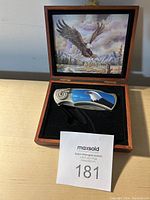 Open wooden box showing folding knife with eagle motif handle and inside lid painting of an eagle in flight over a natural landscape.