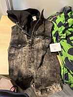 Photo showing black faded denim jacket with buttons and attached hood, lying flat on floor with a foot visible next to it.