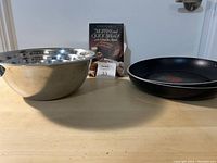 Photo showing two black frying pans, two stainless steel mixing bowls stacked, and a baking book on a kitchen counter.