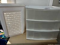 Photo showing a white plastic laundry basket with a grid ventilation pattern and a four-drawer white plastic dresser with transparent drawers, placed next to each other on a wooden surface.