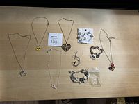 Photo of assorted necklaces and jewelry laid out on wooden surface, showing various pendant shapes including heart, smiley face, clover, and bracelets.