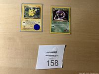 Photo shows two first edition Pokemon cards: Pikachu and Koga's Arbok, placed on a light wood surface with auction lot number 158 nearby.