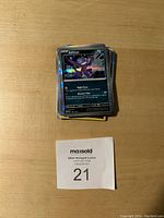 Stack of holographic and regular Pokemon trading cards featuring Sableye on top, arranged on a wooden surface with MaxSold lot number 21 card.