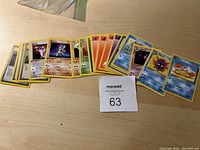 Photo shows about 20 first edition Pokemon cards spread out on a wooden surface with a MaxSold lot number 63 card. Cards feature diverse Pokemon types including Psychic, Fire, Water, and Energy cards, all with the first edition symbol and clear artwork.