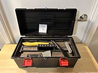 Photo of an open black plastic toolbox containing various hand tools including a wire brush, scrapers, utility knife, metal file, and a triangular metal tool.