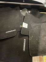 Top view of four carpet floor mats in black with Corolla logo, showing trimmed edges and minor use wear