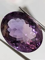 Close-up image of large oval cut faceted amethyst gemstone held by tweezers, showing rich purple color and clarity.