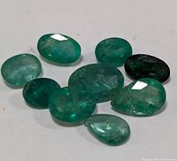Nine loose genuine emerald gemstones arranged on white background, varying in size and shape, faceted with visible inclusions and color variations.