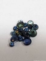 Close-up photo of a pile of various loose sapphire gemstones in blue and green hues on a white background.