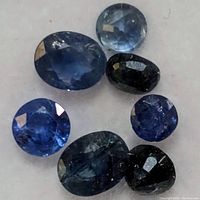 Seven loose genuine sapphire gemstones in assorted oval and round faceted cuts and various blue colors, displayed on a white background.