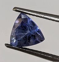 Close-up photo of a triangular faceted blue-violet tanzanite gemstone held by tweezers, showing surface facets and natural inclusions.
