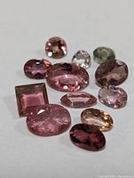 Loose faceted assorted tourmaline gemstones in various shapes and shades mostly pink with one light green stone on white background.