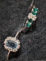 Close-up of two silver rings: one with a rectangular emerald-cut blue stone encircled by small clear stones, the other with green and clear stones arranged in clusters on a silver band.