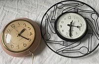 Two kitchen clocks placed on a white cloth, the electric clock is pink with gold trim, the battery clock black with cutlery design frame.