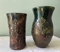 Two pottery vases side by side on a white cloth with a light blue background; one vase is cylindrical with dark brown iridescent raised flower patterns, the other is taller, dark green with iridescent flower patterns and a flared rim.