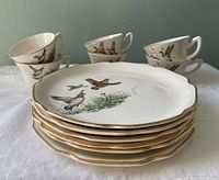Stack of six luncheon plates with scalloped edges and gold trim, each decorated with different bird scenes. Six matching cups arranged behind plates showing varied bird patterns.