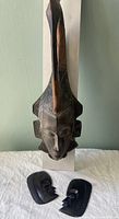 Tall African wooden mask carved with a bird headpiece mounted on a white display board, viewed frontally with two face plaques positioned below.
